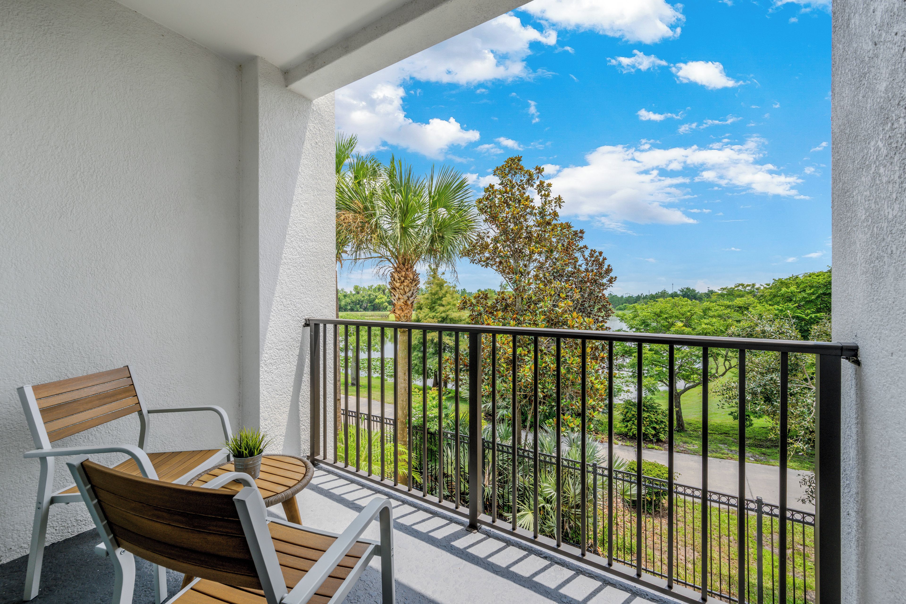 A balcony with a wooden table and chairs, a small potted plant, and a view of a green landscape with trees and a clear sky.