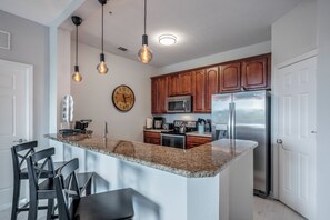 A modern kitchen with granite countertops, stainless steel appliances, and wooden cabinets. There is a large island with bar stools, and pendant lighting hangs above. - Is1-208 - 3BR Condo Near Disney With Lake View (Orlando)