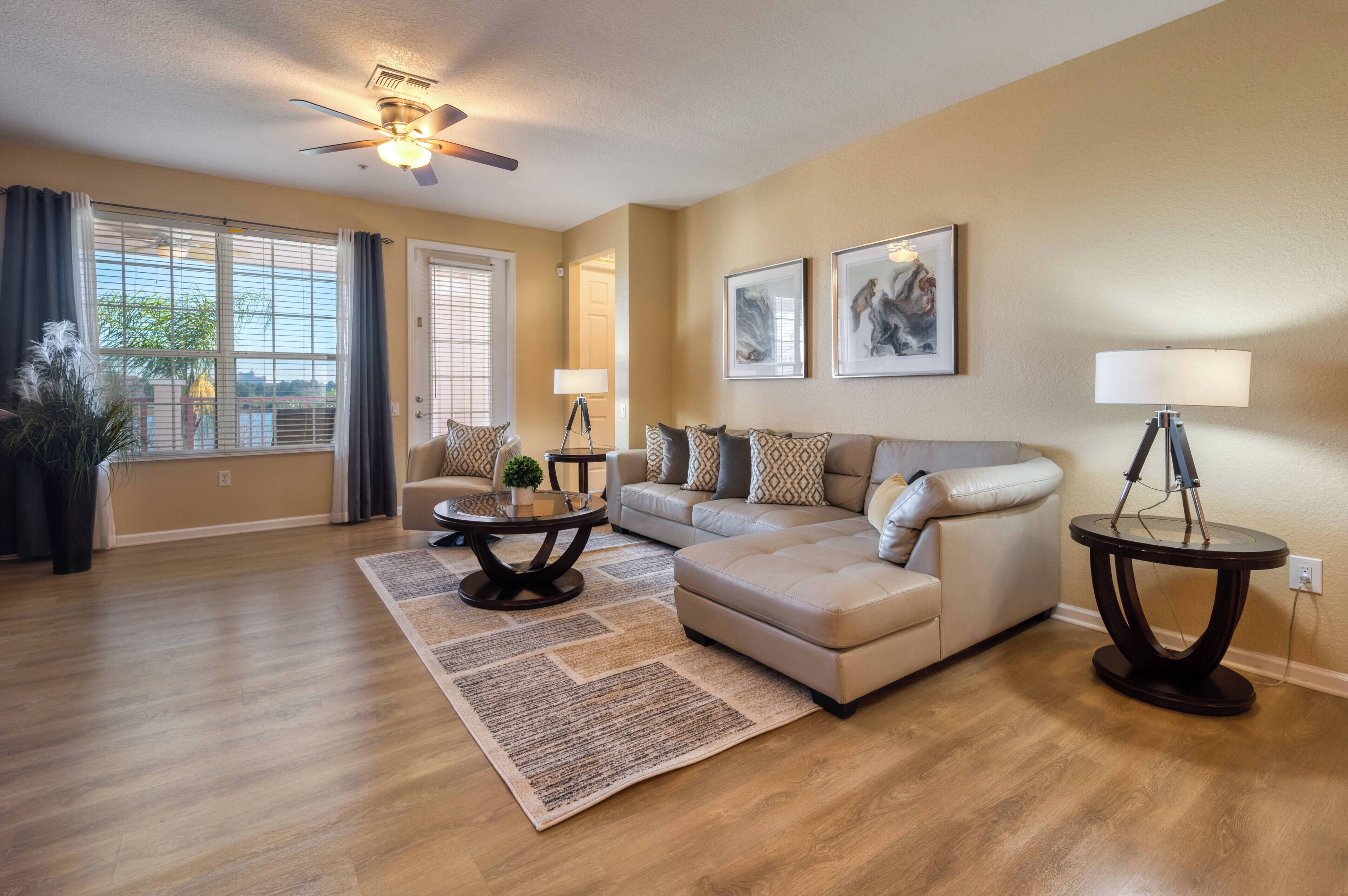 Un salon avec un canapé beige, une table basse et un lampadaire. Deux tableaux encadrés ornent le mur, et un ventilateur de plafond est installé au plafond.