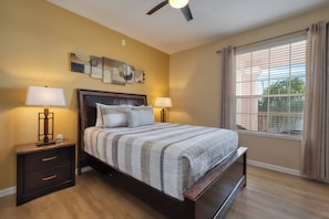 Une chambre à coucher comprenant un lit, deux tables de chevet, un ventilateur de plafond et une fenêtre avec des rideaux.