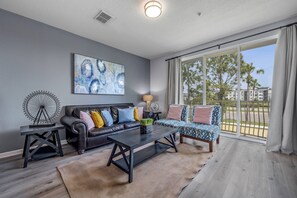 A modern living room with a dark leather sofa, a patterned sofa, and a coffee table. There is a large window with a view of buildings and trees. - Th155 - 3BR House Near Disney, Sleeps 8 Guests (Orlando)