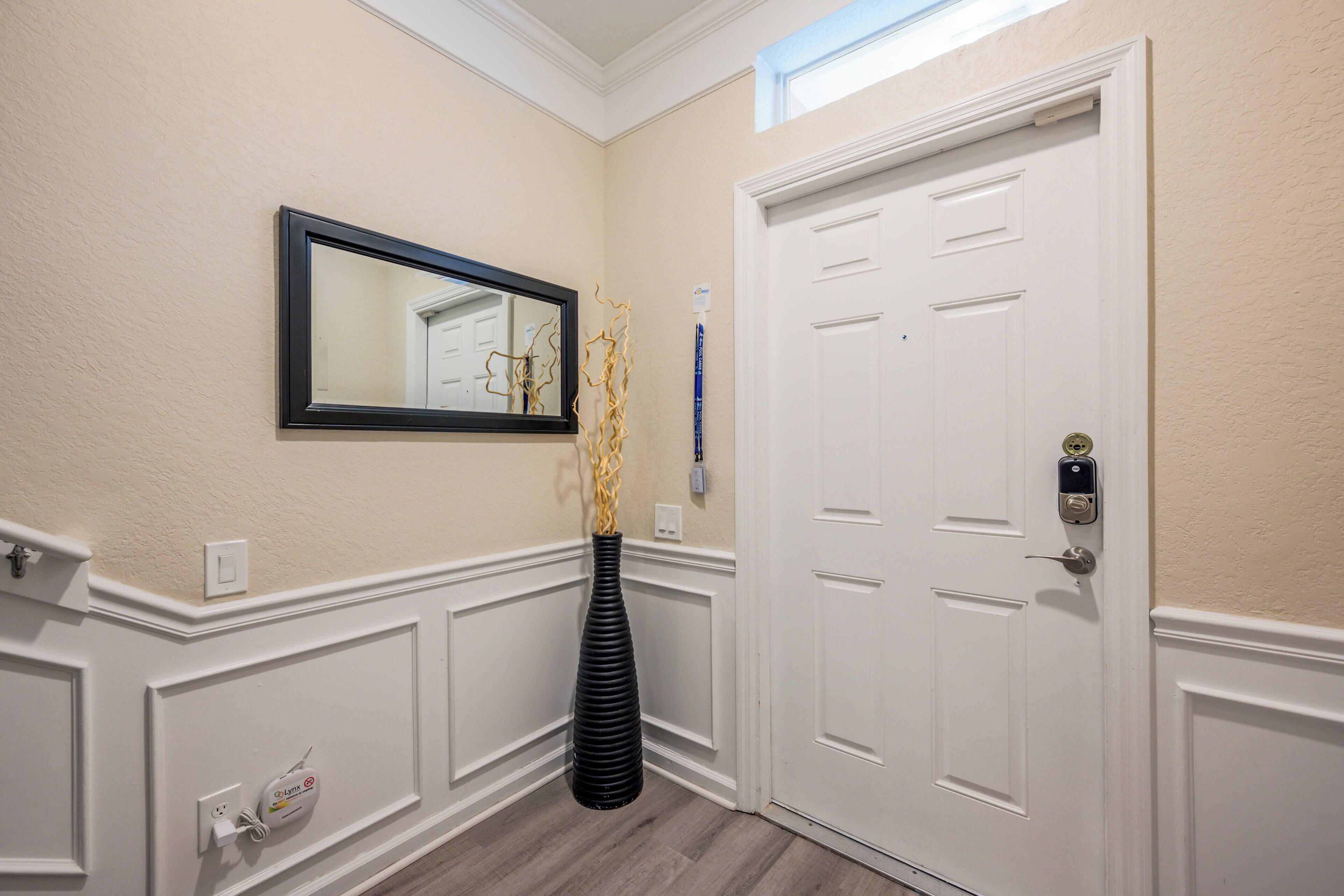 Un pasillo con una puerta blanca, un espejo enmarcado y un jarrón negro con ramas decorativas.