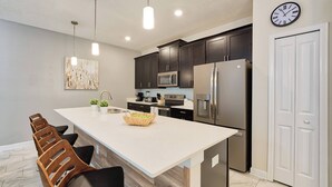 A modern kitchen with dark wooden cabinets, stainless steel appliances, a large island with bar stools, and a wall clock. - Sl4926 - 4BR House With Pool Near Disney Area (Kissimmee)