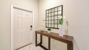 A hallway with a white door, a wooden console table with a vase and a plant, and a framed mirror on the wall. - Sl4926 - 4BR House With Pool Near Disney Area (Kissimmee)