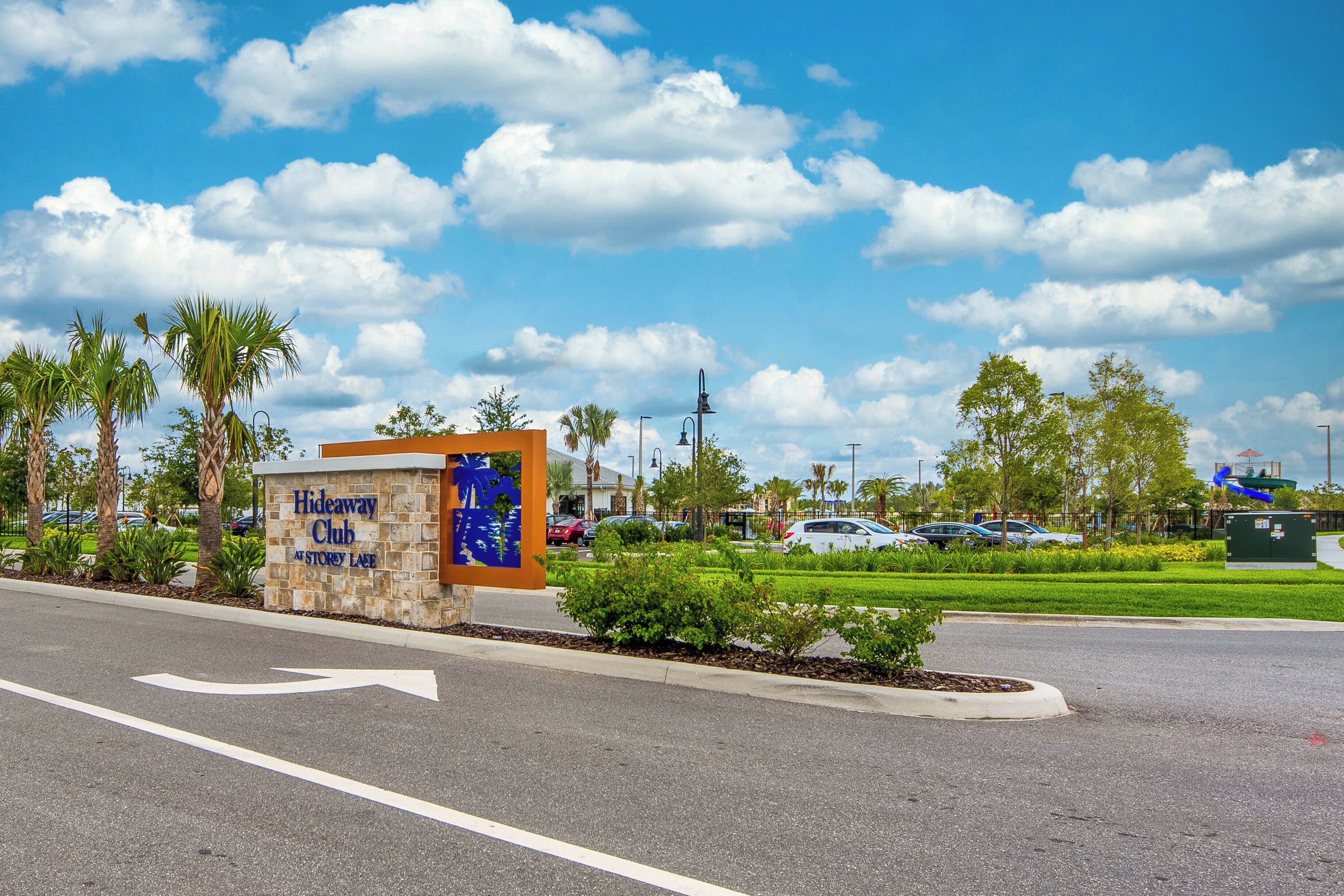 A sign for the Hideaway Club at Story Lake with a road leading to it, surrounded by palm trees and a clear sky.