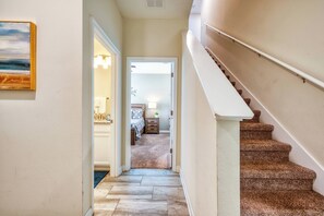Un couloir menant à une chambre dont le sol est recouvert d’une moquette, avec un escalier et un tableau accroché au mur.