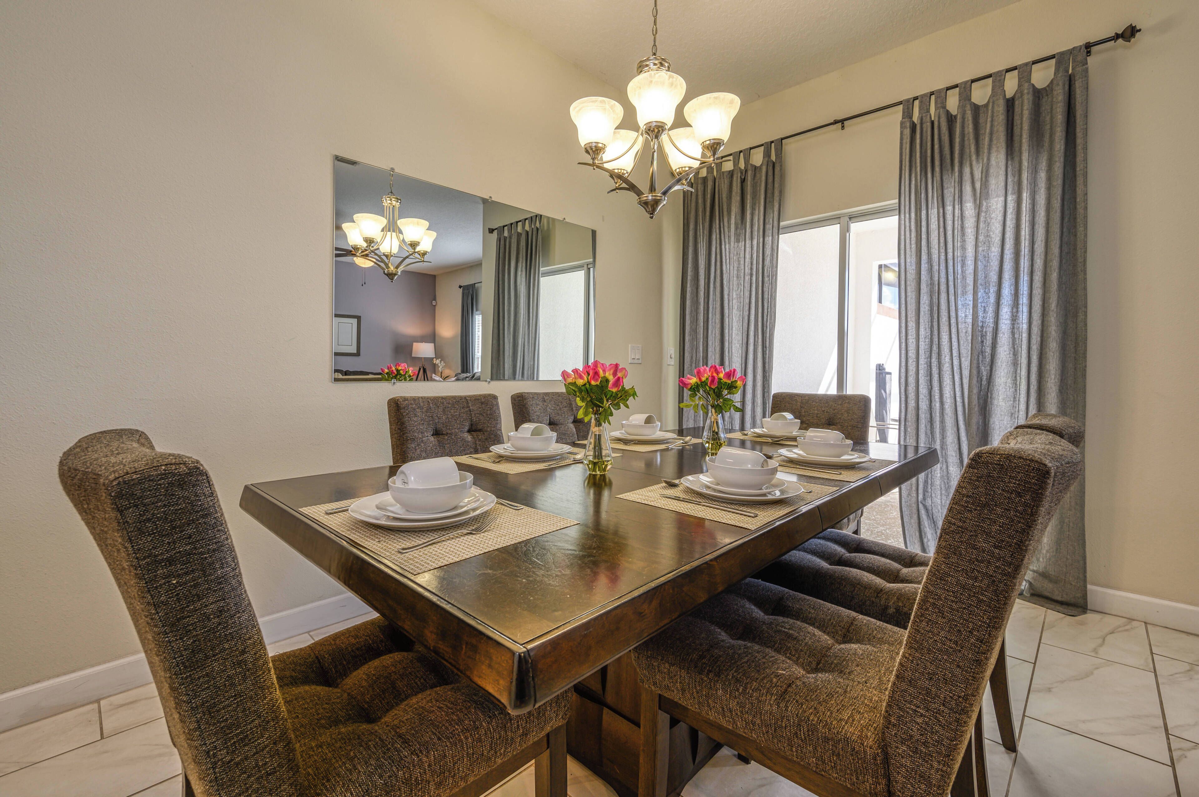 A dining area with a long table set for six, surrounded by upholstered chairs, a chandelier, and a large mirror above the table.
