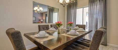 A dining area with a long table set for six, surrounded by upholstered chairs, a chandelier, and a large mirror above the table.
