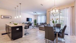 Eine moderne Küche mit Kochinsel, einem Essbereich mit Tisch und Stühlen, einem an der Wand montierten Flachbildfernseher und einem großen Fenster mit Vorhängen.
