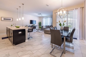 A modern kitchen with a central island, a dining area with a table and chairs, a flat-screen TV mounted on the wall, and a large window with curtains. - Sl4924 - 4BR House With Pool Near Disney (Kissimmee)