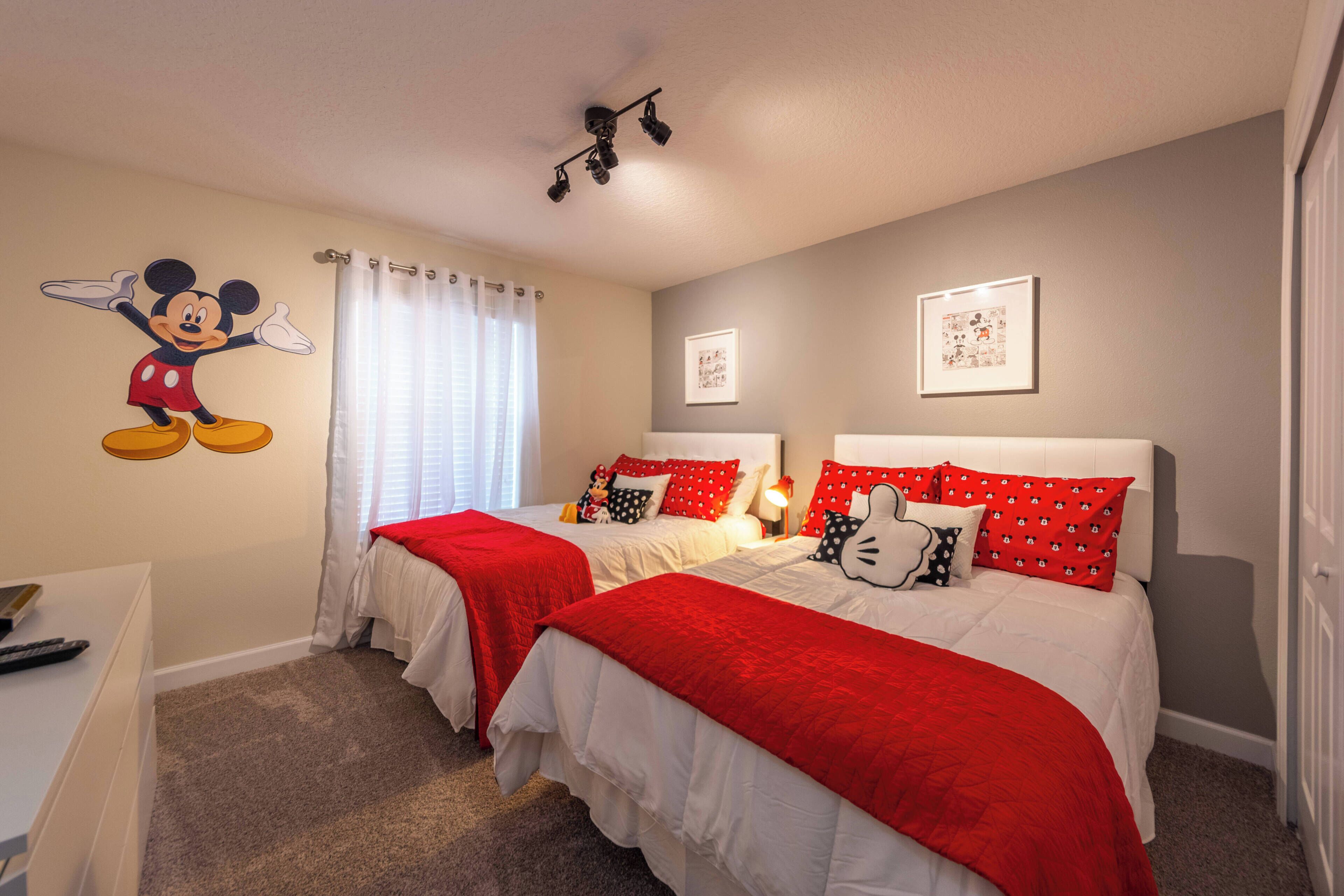 A hotel room with two beds, red and white bedding, a Mickey Mouse wall decal, and two framed pictures on the wall.