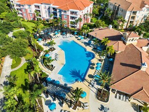 Vista aérea de um resort com uma piscina central, várias áreas de estar com guarda-sóis e diversos edifícios com telhados de telha vermelha.