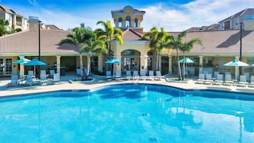 A swimming pool with lounge chairs and umbrellas, surrounded by palm trees and buildings with tiled roofs.