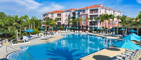 A hotel with a large pool surrounded by lounge chairs and umbrellas, palm trees, and a clear blue sky.