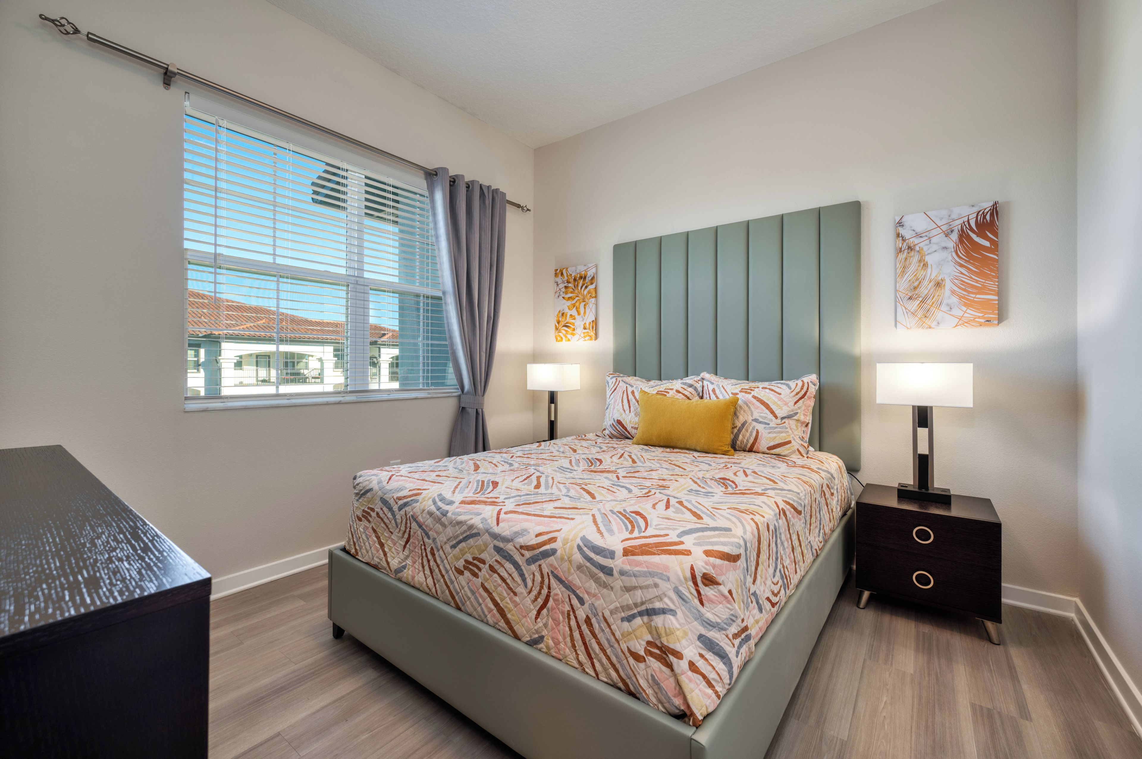 A modern bedroom with a bed, two bedside tables, a nightstand, a window with curtains, and artwork on the walls.