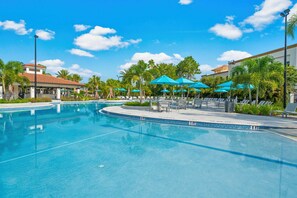 A swimming pool with lounge chairs and umbrellas, surrounded by palm trees and a building with a red roof. - Vcr1-207 - 3BR Condo Near Disney at Vista cay (Orlando)