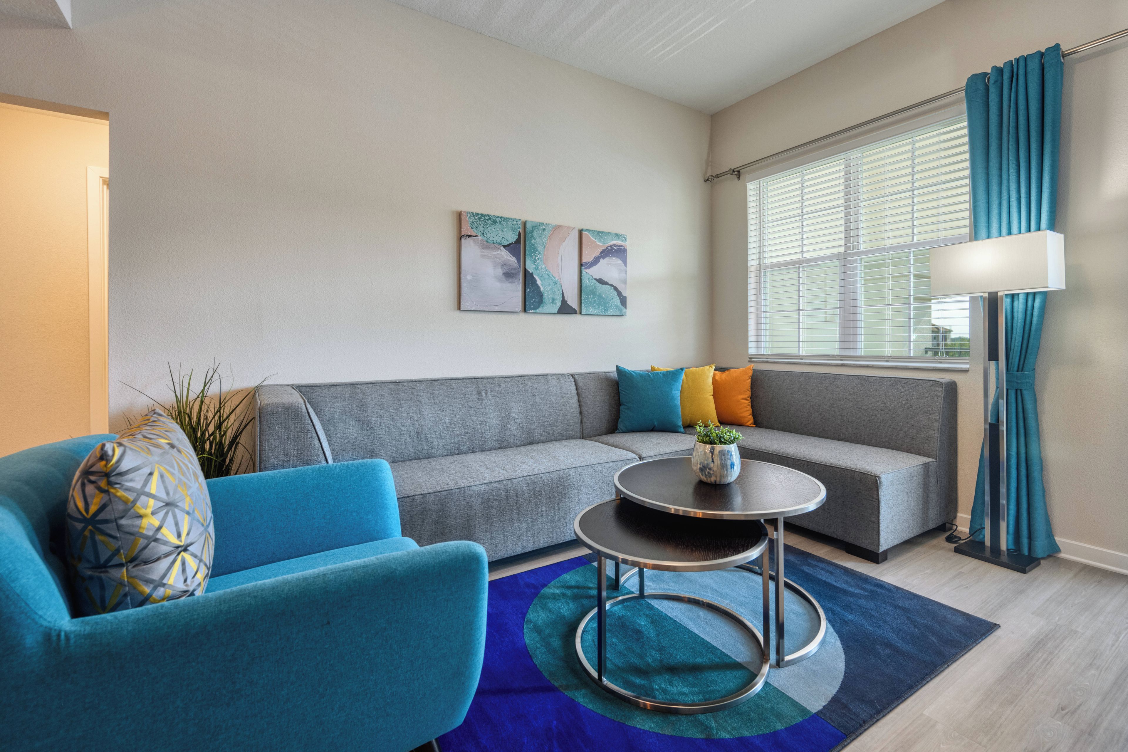 Een moderne woonkamer met een grijze bank, een blauwe fauteuil en een ronde salontafel. Aan de muur hangen abstracte schilderijen en er is een raam met gordijnen.