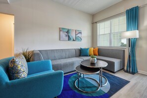 Una sala moderna con un sofá gris, un sillón azul y una mesa de centro redonda. Hay pinturas abstractas en la pared y una ventana con cortinas.