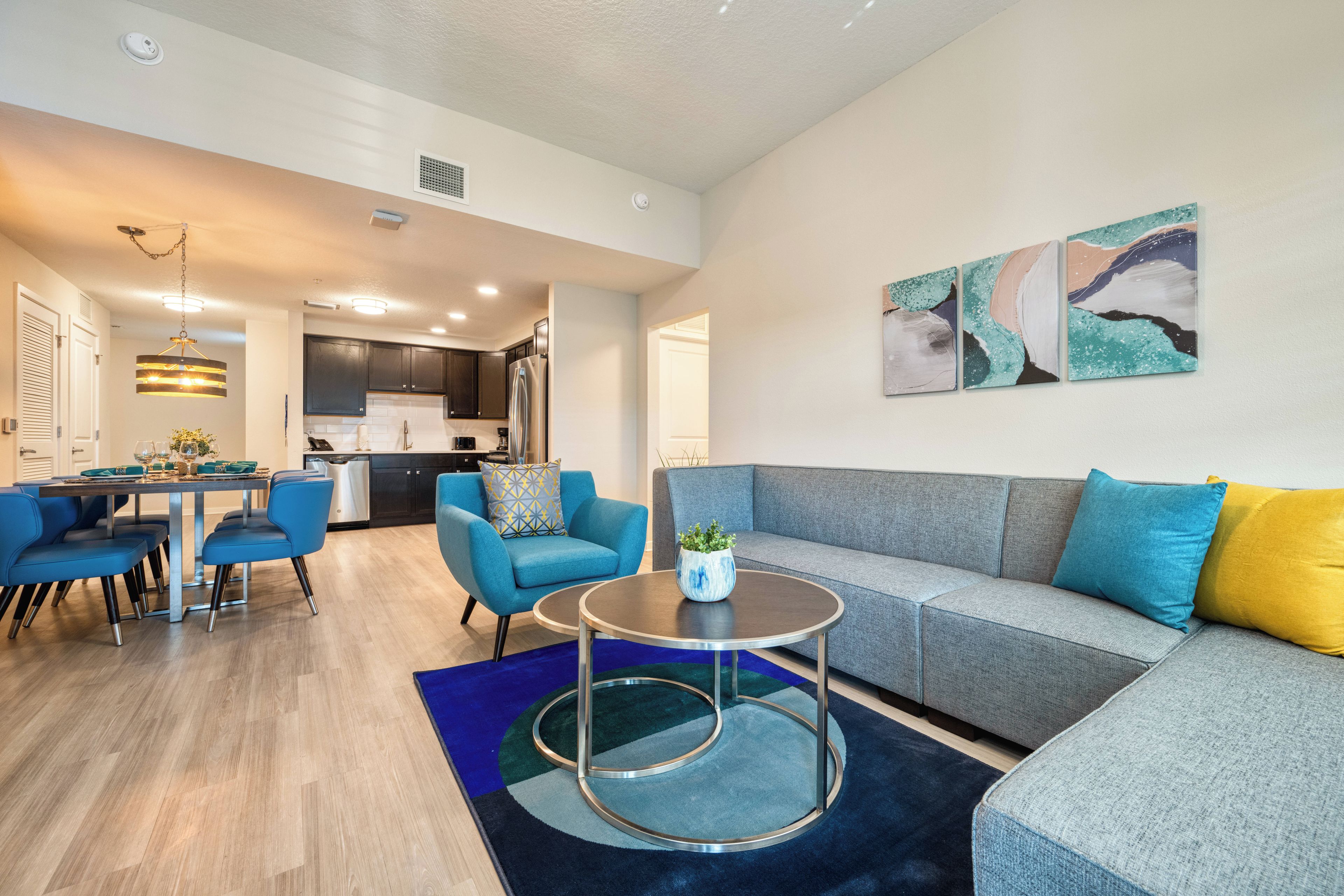 Un salon moderne avec un canapé gris, des fauteuils bleus, une table basse ronde et un coin repas comprenant une table et des chaises.