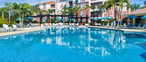 A clear blue pool with lounge chairs and umbrellas, surrounded by palm trees and a building with balconies.