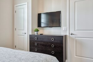 Un dormitorio con una cama, un aparador de madera oscura, un televisor de pantalla plana montado en la pared y una pequeña planta en maceta sobre el aparador.