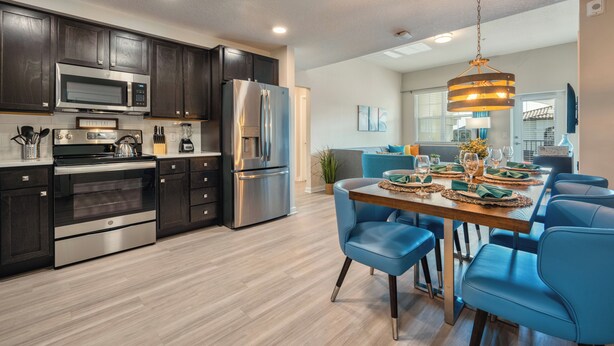 A modern kitchen with dark cabinets, stainless steel appliances, and a dining area with blue chairs and a wooden table. - Vcr4-410- 3BR Condo With Resort Access Near Disney (Orlando)