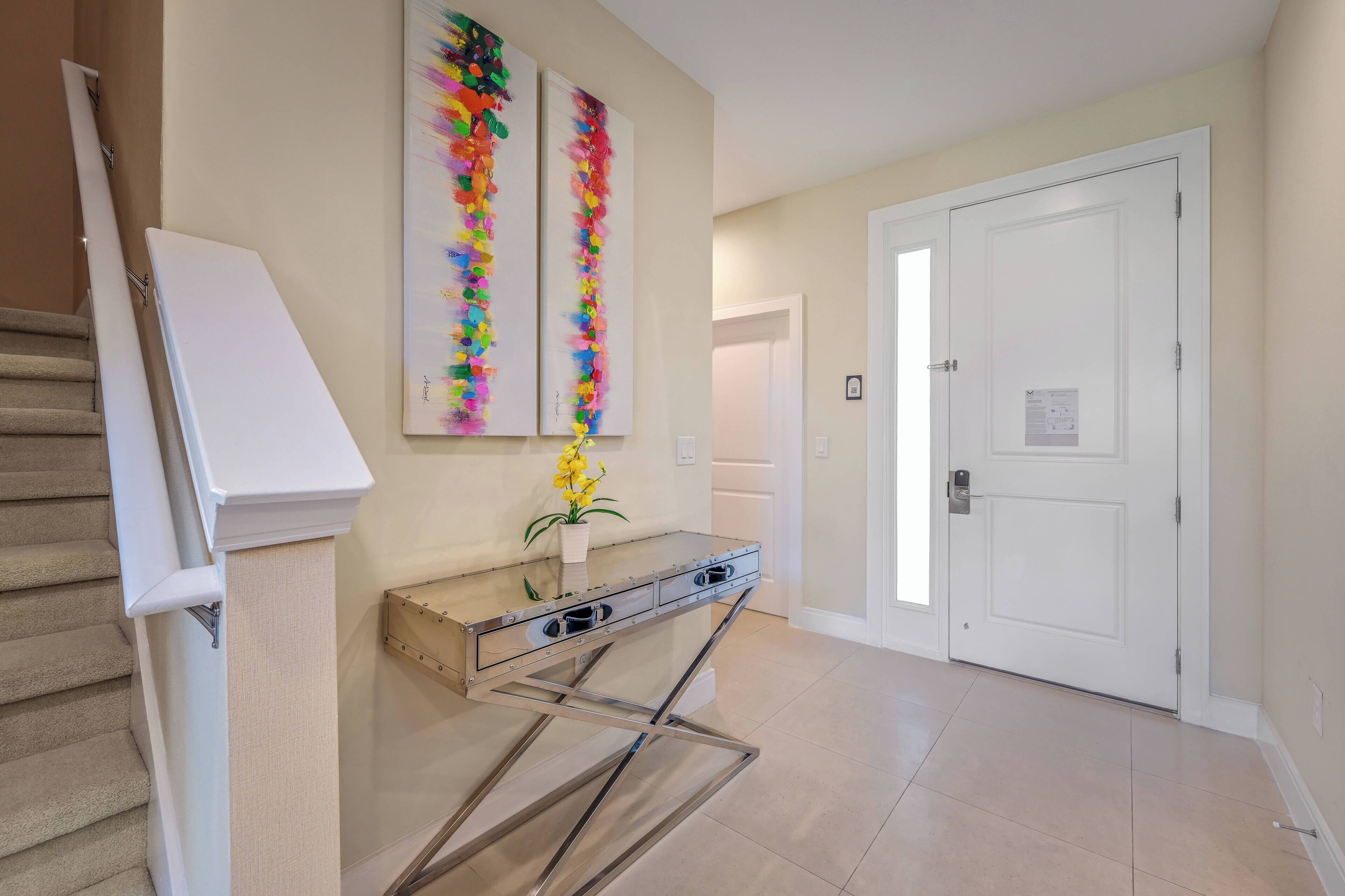 Un interior moderno con una escalera, una consola de vidrio con cajones y dos cuadros abstractos en la pared.