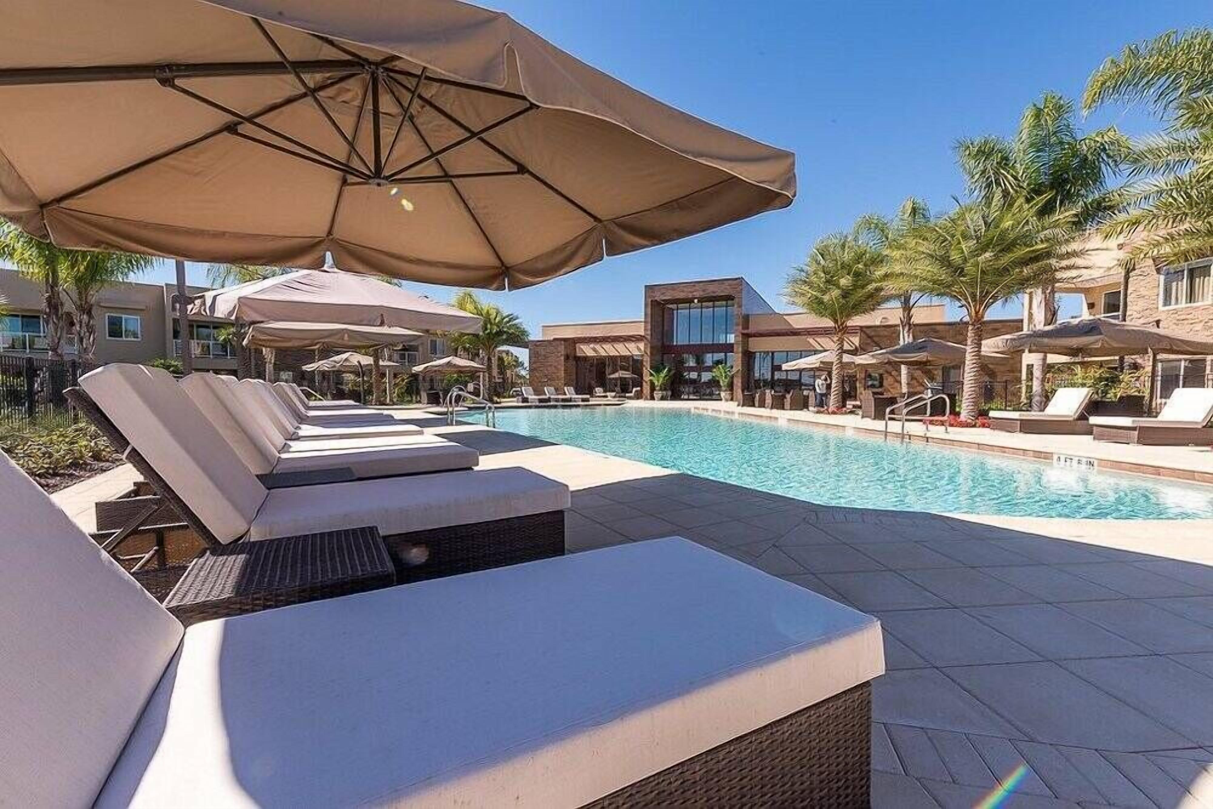 Zona junto a la piscina con sillas de descanso y sombrillas, rodeada de palmeras y edificios modernos.