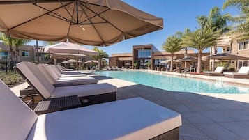 Zona junto a la piscina con sillas de descanso y sombrillas, rodeada de palmeras y edificios modernos.