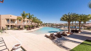 A poolside area with lounge chairs and palm trees, a clear blue sky, and a building with a modern design. - Mvy7664 - 3BR House Near Disney With Hot Tub (Kissimmee)