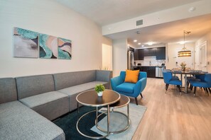 A modern living room with a grey sectional sofa, a blue armchair, a round coffee table, and a dining area with blue chairs and a chandelier. - Vcr4-304 3BR Condo With Resort Access Vista Cay (Orlando)