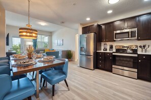 A modern kitchen with stainless steel appliances, dark wooden cabinets, and a dining area with blue chairs and a wooden table set for a meal. - Vcr4-304 3BR Condo With Resort Access Vista Cay (Orlando)