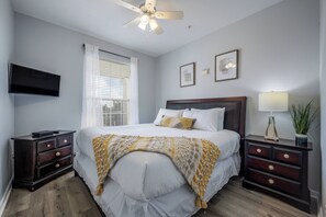 Ein Schlafzimmer mit Bett, Nachttischen, einem Fernseher, einem Deckenventilator und einem Fenster mit Jalousien.
