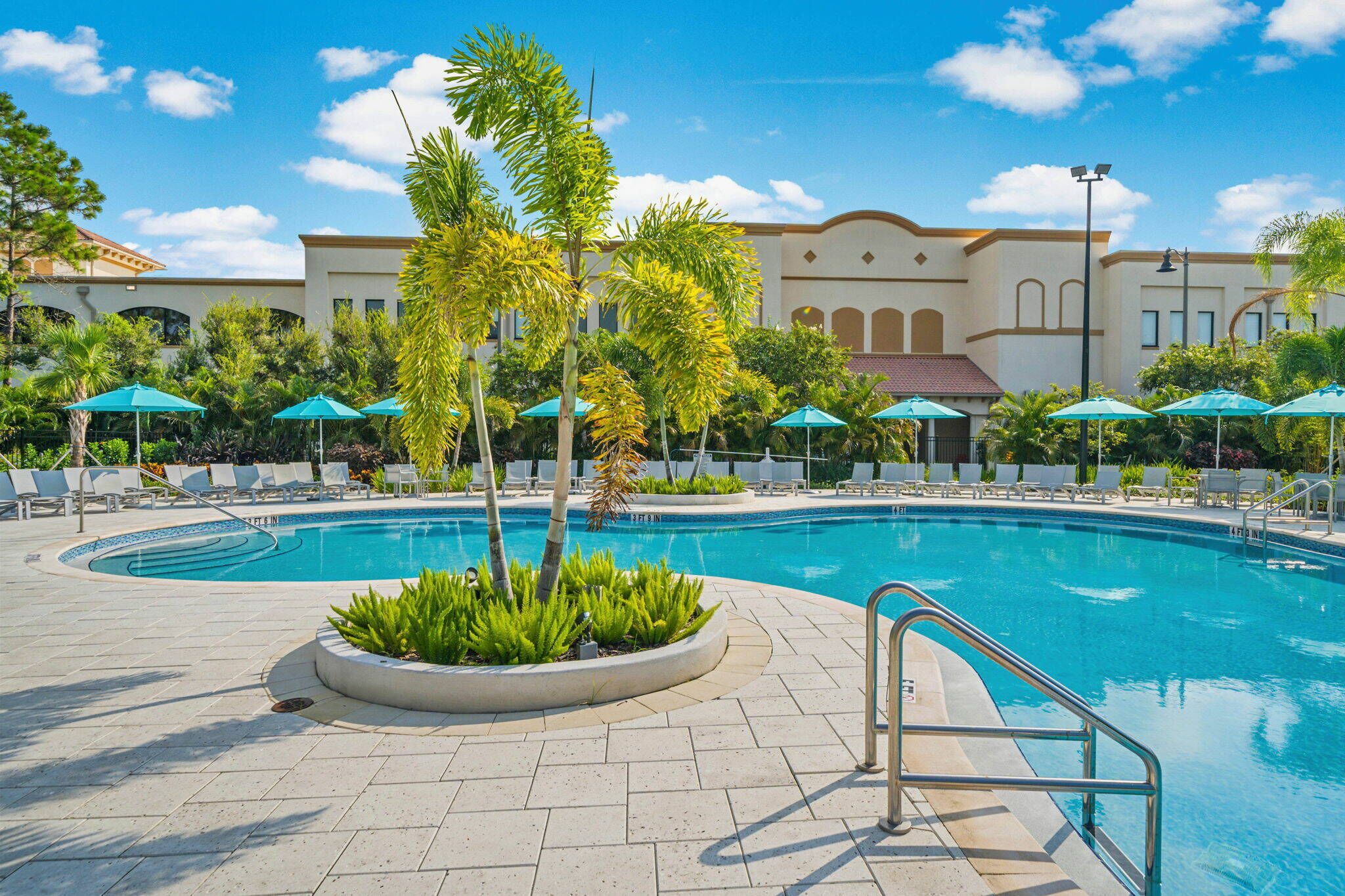 A swimming pool with lounge chairs and umbrellas, surrounded by palm trees and a building with arched windows.