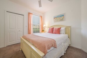 Un dormitorio con cama, mesita de noche, ventana con cortinas, ventilador de techo y un cuadro en la pared.