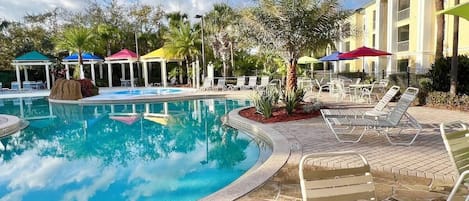 A swimming pool with lounge chairs and umbrellas, surrounded by palm trees and a building with multiple windows.