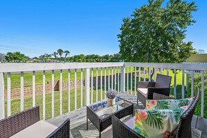 A deck with wicker furniture and a glass table, overlooking a grassy area with a white fence and trees. - Sw40 - 3BR Apartment Near Disney With Resort Pool (Kissimmee)