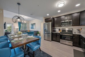 Una cocina moderna con armarios oscuros, electrodomésticos de acero inoxidable y un comedor con sillas azules y una mesa de madera dispuesta para seis comensales.