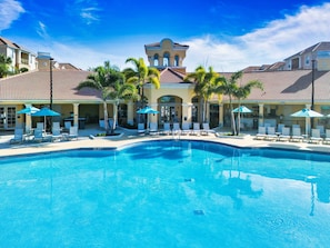 A swimming pool with lounge chairs and umbrellas, surrounded by palm trees and buildings with tiled roofs. - Vcr1-104 - 3BR Condo at Vista Cay Resort (Orlando)