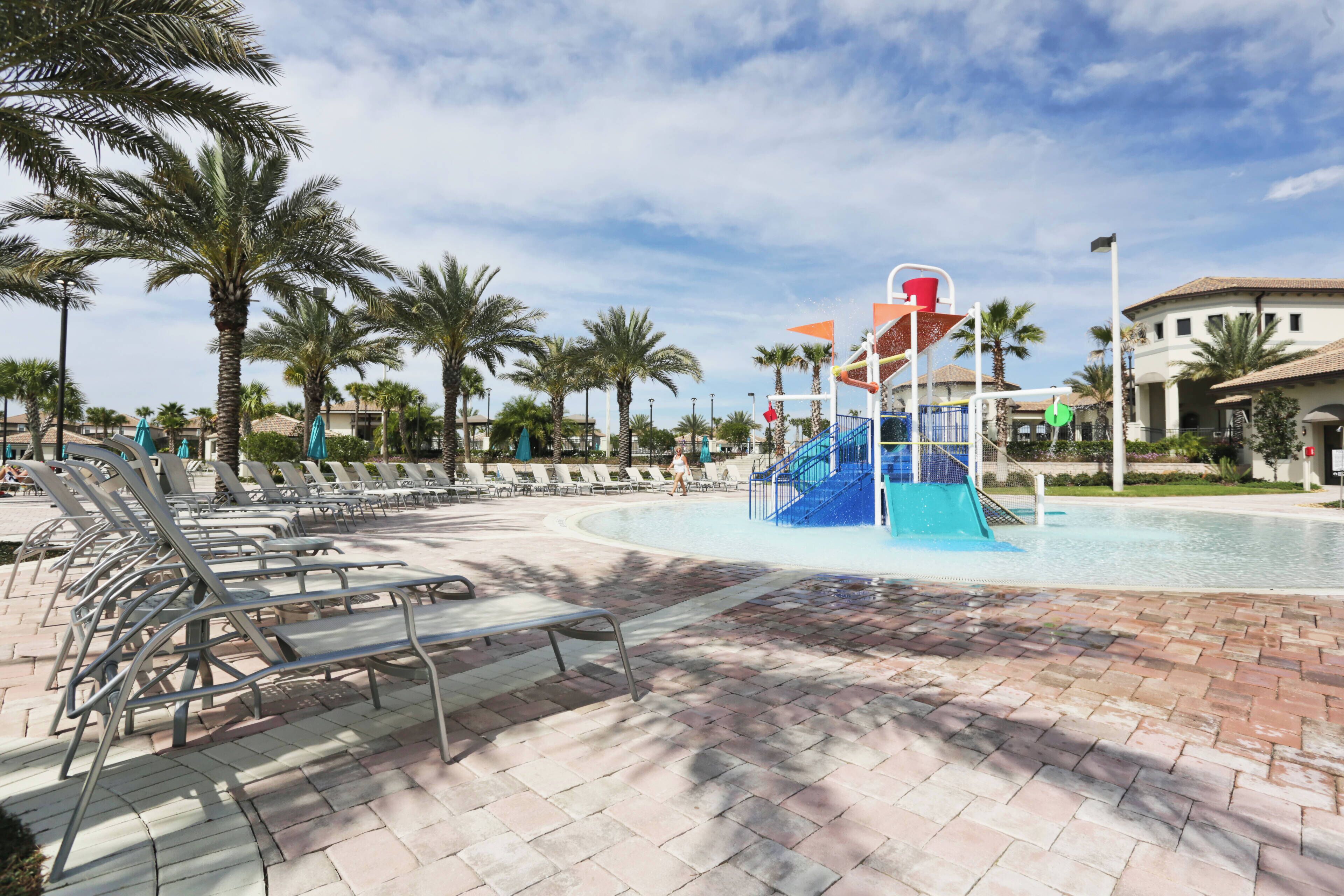 A pool area with lounge chairs, a water slide, palm trees, and a building in the background.