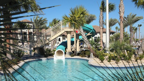 A swimming pool with a water slide, palm trees, and a building with a red roof in the background. - Cg1569 - 5BR House With Pool Near Disney Parks (Davenport)