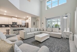 A spacious living area with a large sectional sofa, a coffee table, and a kitchen in the background. - Cg1569 - 5BR House With Pool Near Disney Parks (Davenport)