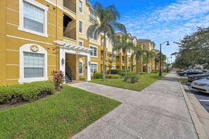 A yellow multi-story residential building with a covered entrance, palm trees, and a sidewalk. - Vc13-303 - 3BR Condominium Near Parks (Orlando)