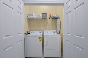 A laundry room with two white washers and dryers, a shelf with towels and laundry supplies, and a hanging rack with clothes. - Vc13-303 - 3BR Condominium Near Parks (Orlando)