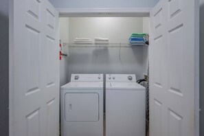 A laundry room with two white front-loading washers and dryers, a metal shelf above for folded clothes, and a white door with a keyhole lock. - Vc14-407 - 2BR Epic Condo Near Disney (Orlando)