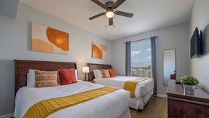 Habitación de hotel con dos camas, ventilador de techo, obras de arte en las paredes, ventana con cortinas, televisor montado y cómoda con una planta.