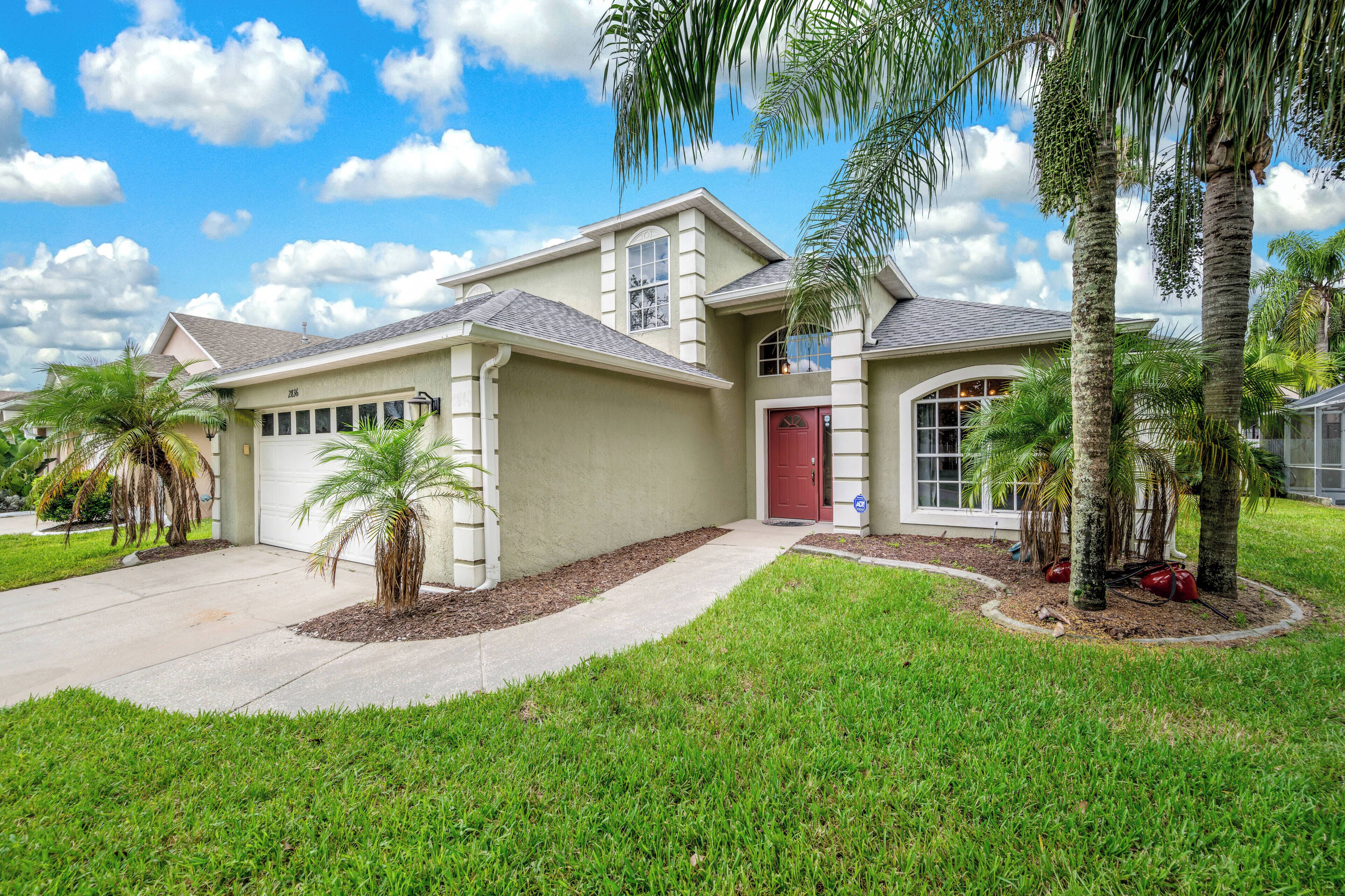 A two-story house with a garage, a red door, and a well-maintained lawn with palm trees.