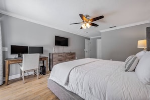 Ein Schlafzimmer mit einem großen Bett, einem Schreibtisch mit zwei Bildschirmen, einem Stuhl, einem Deckenventilator und einem Fernseher an der Wand.