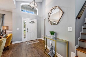 A modern entryway with a wooden floor, a white door, a glass console table with a plant, and a geometric mirror on the wall. - Oi2836 - 4BR House With Pool Near Disney World (Kissimmee)