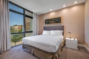 A modern hotel room with a large bed, a nightstand, a wall-mounted artwork, and a view of the outdoors. - Mvv7482 - 3BR House Near Disney at Magic Village (Kissimmee)